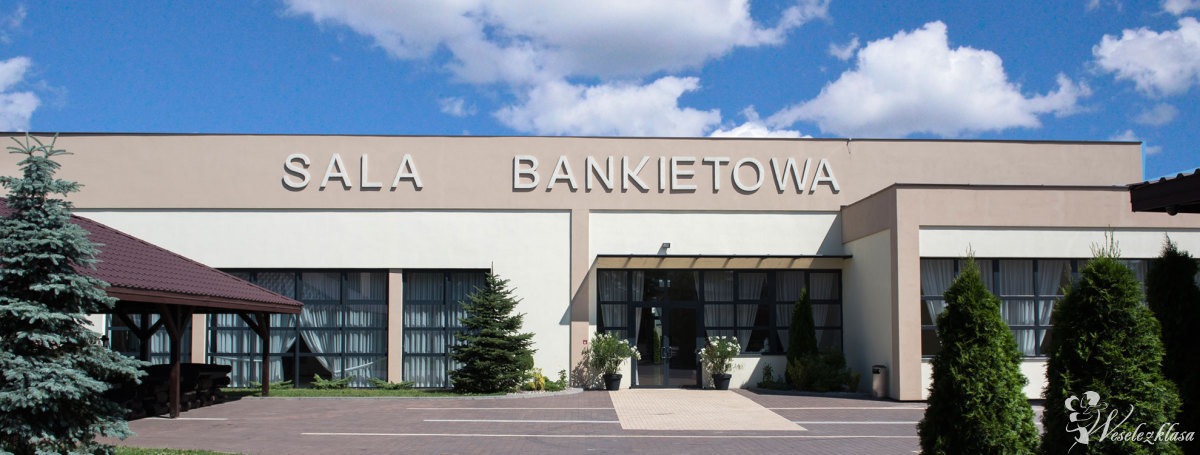 Elewacja budynku z napisem 'SALA BANKIETOWA' w słoneczny dzień, z widocznymi drzewami iglastymi i zadaszoną wiatą.