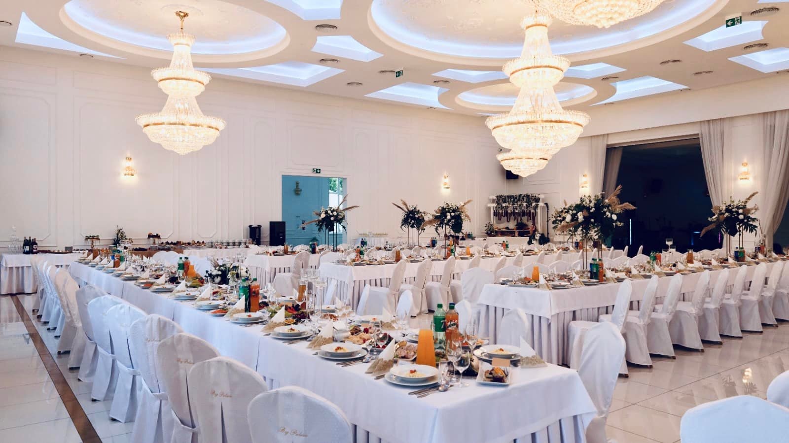 Elegancka sala bankietowa z długimi stołami nakrytymi białymi obrusami, krzesła w pokrowcach, dekoracje kwiatowe i kryształowe żyrandole