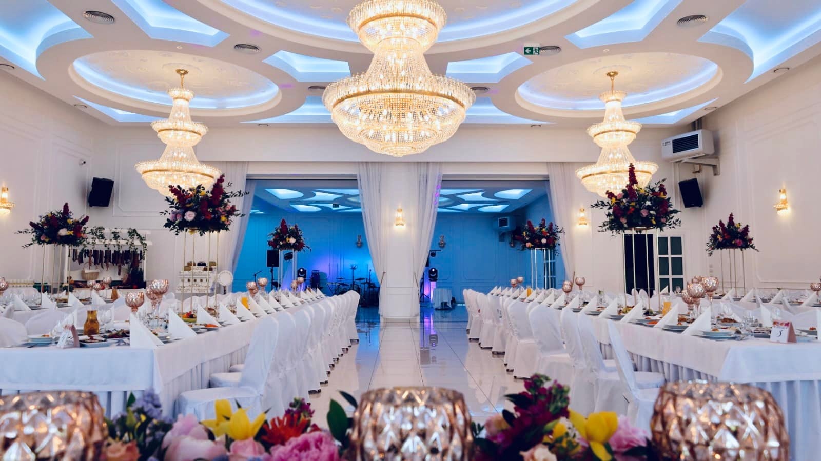 Uroczysta sala bankietowa z długimi stołami nakrytymi białymi obrusami, krzesła w pokrowcach, kryształowe żyrandole, dekoracje kwiatowe i oświetlenie sceniczne.