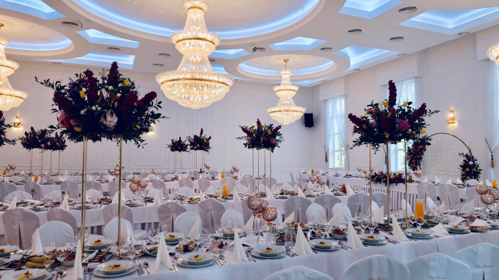 Uroczysta sala bankietowa z długimi stołami nakrytymi białymi obrusami, dekoracjami kwiatowymi na wysokich złotych stojakach, kryształowymi żyrandolami i dekoracyjnym oświetleniem sufitowym.