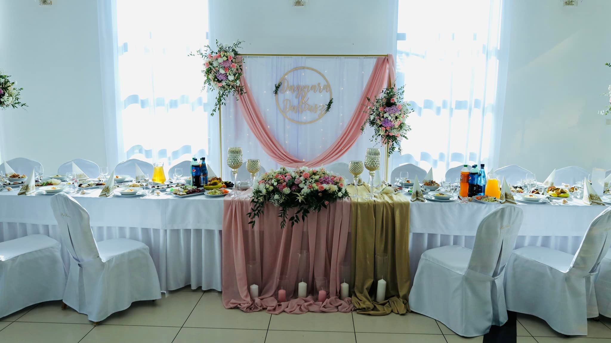 Uroczysty stół weselny z białym obrusem, dekoracjami kwiatowymi w odcieniach różu i fioletu, złotymi świecznikami i napojami w tle białych zasłon.