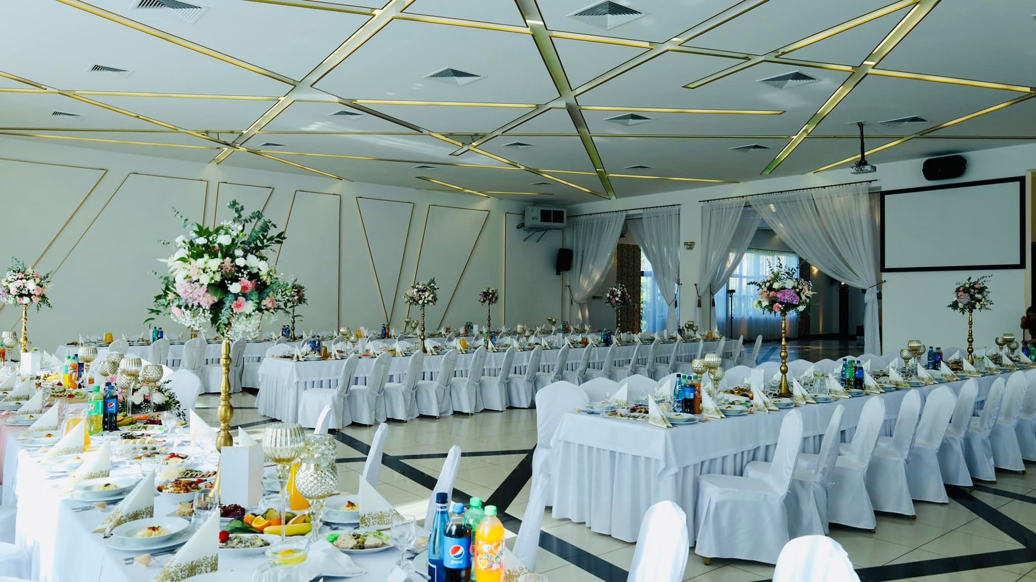 Uroczysta sala bankietowa z długimi stołami nakrytymi białymi obrusami, krzesła w pokrowcach, dekoracje kwiatowe w złotych wazonach, zastawa stołowa i napoje, ściany zdobione złotymi detalami.