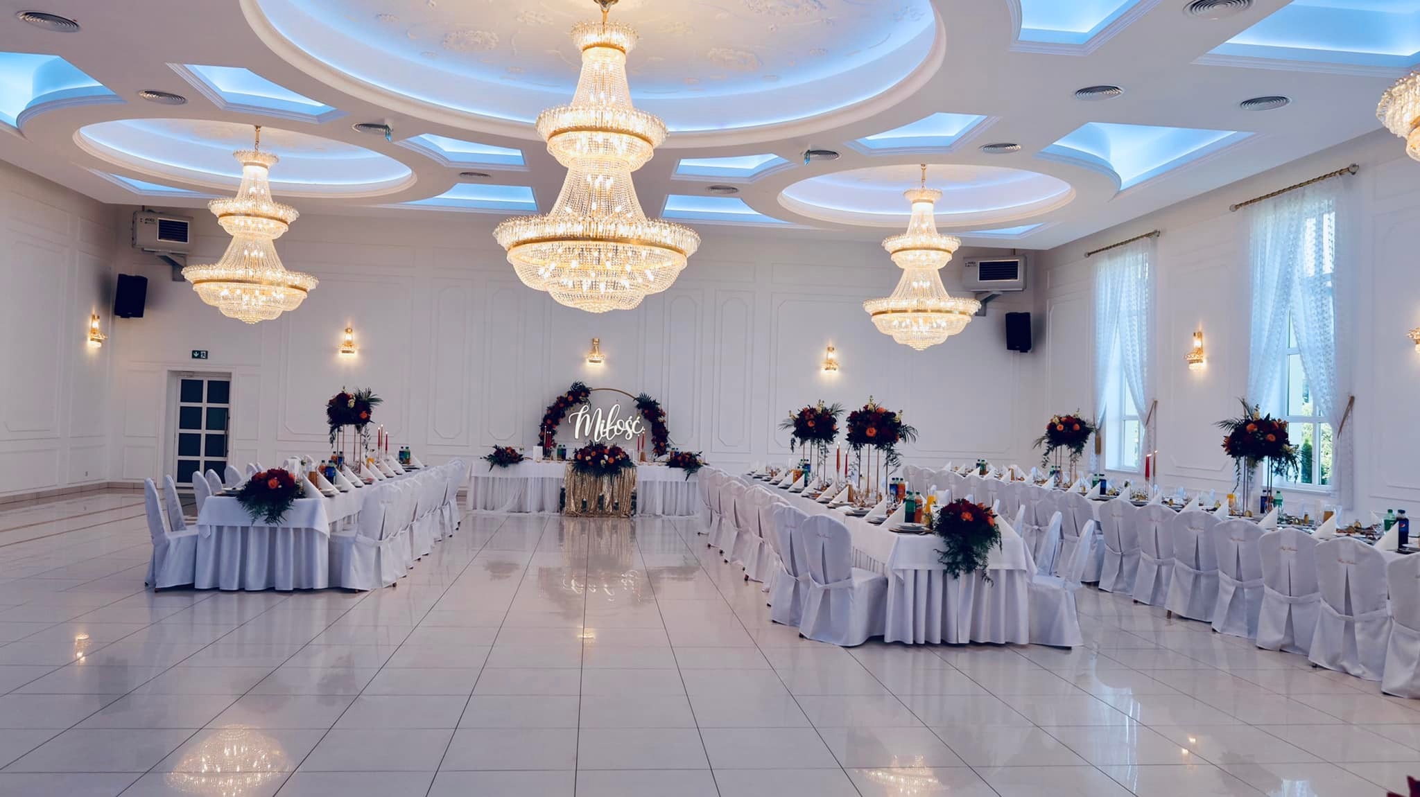 Sala bankietowa z białymi obrusami, krzesłami w pokrowcach, kwiatowymi dekoracjami, kryształowymi żyrandolami i napisem 'Miłość' w tle.