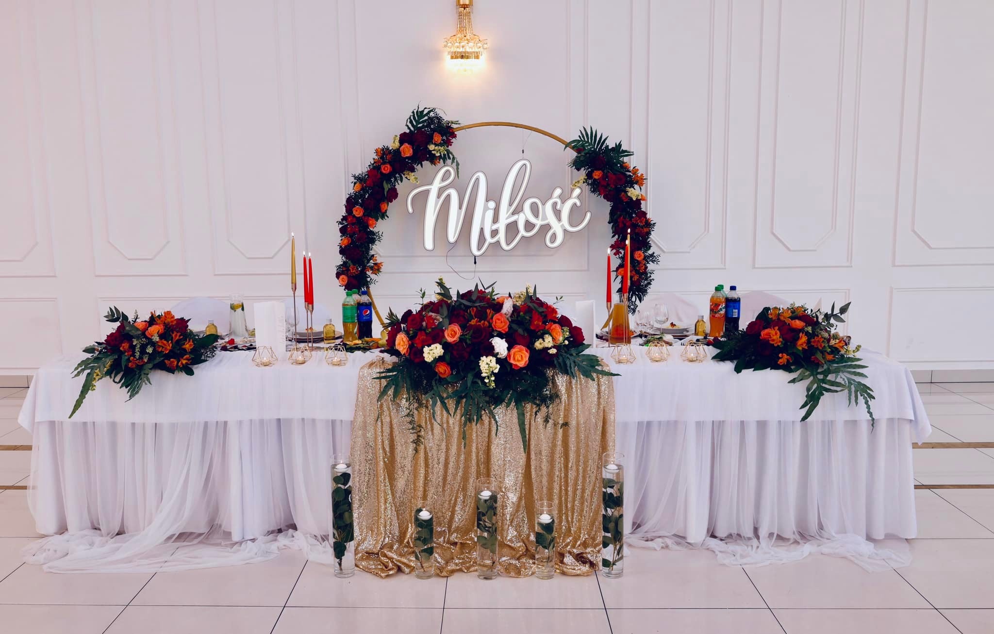 Uroczysta dekoracja stołu weselnego z kwiatami w odcieniach czerwieni i pomarańczy, złotym bieżnikiem z cekinami, świecami i napisem 'Miłość' w okręgu z kwiatów.