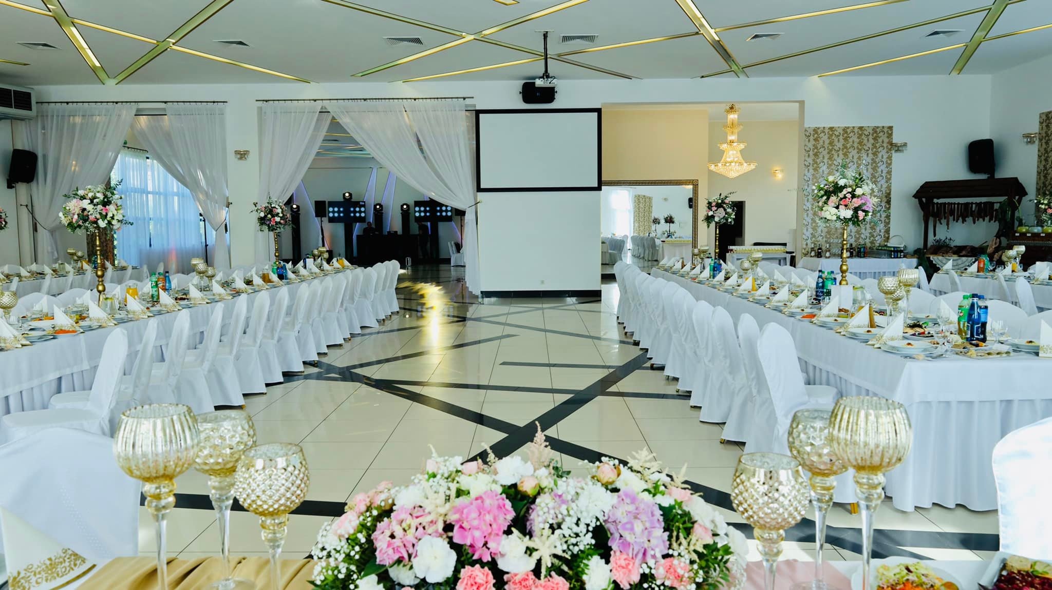 Uroczysta sala balowa z długimi stołami nakrytymi białymi obrusami, białymi krzesłami, kwiatowymi dekoracjami i złotymi świecznikami; widoczny parkiet w czarno-białą szachownicę i scena z projektorem.