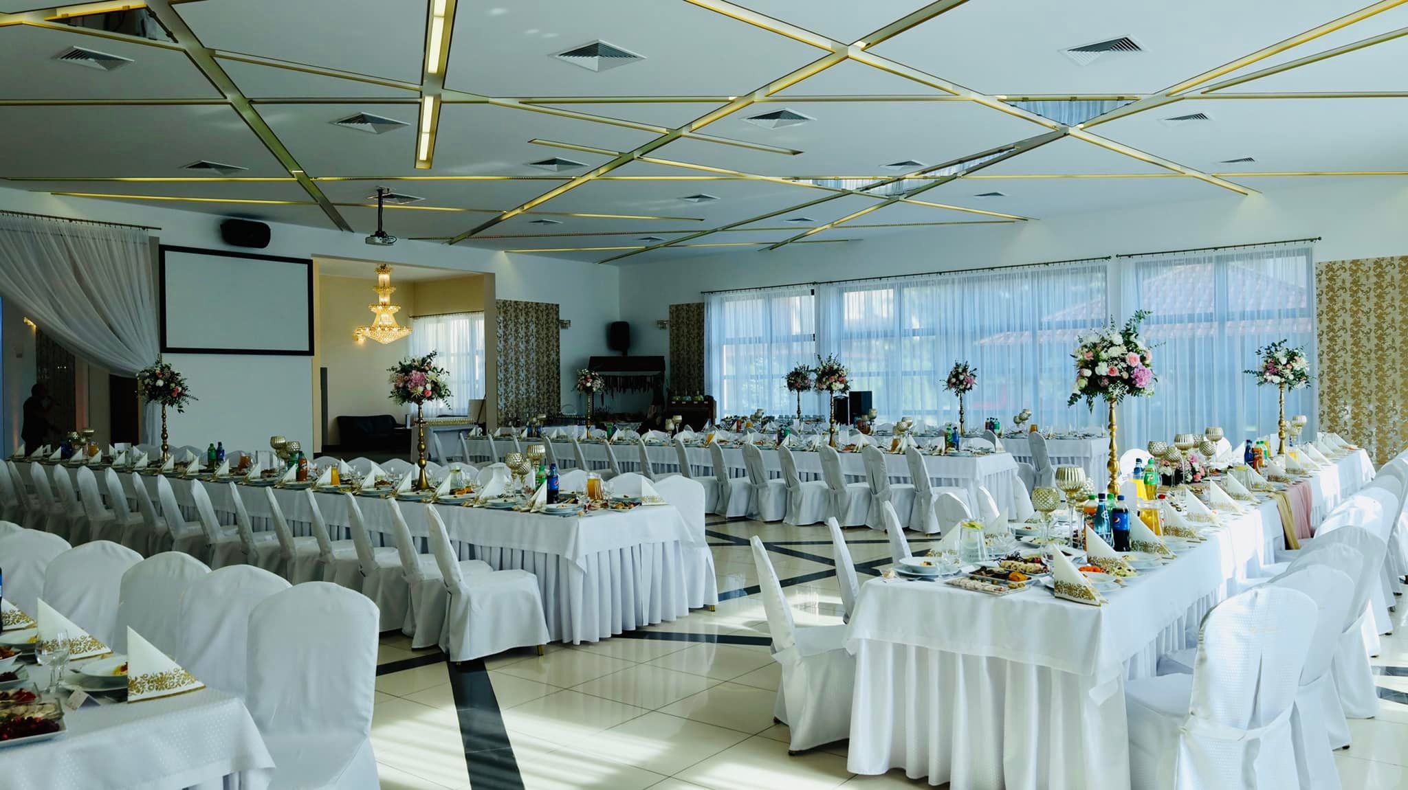 Sala bankietowa z długimi stołami nakrytymi białymi obrusami i krzesłami w pokrowcach, udekorowana kompozycjami kwiatowymi na złotych stojakach, widoczne szklane naczynia, napoje i przekąski...