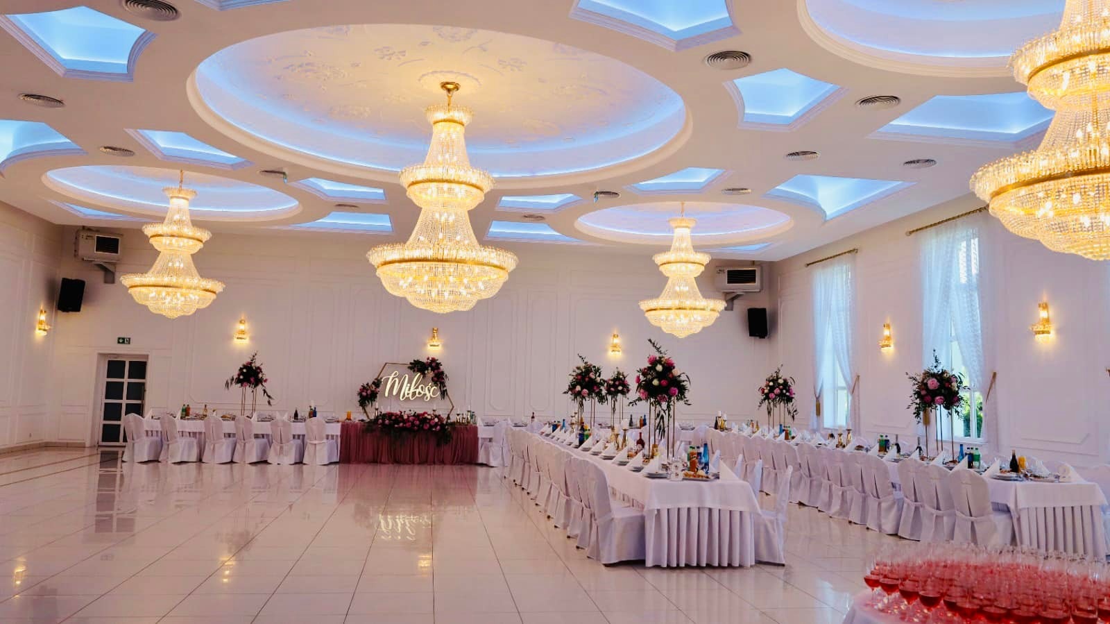 Sala bankietowa z długimi stołami nakrytymi białymi obrusami, dekoracjami kwiatowymi i kryształowymi żyrandolami podświetlonymi niebieskim światłem, napis 'Miłość' na dekoracyjnym tle.