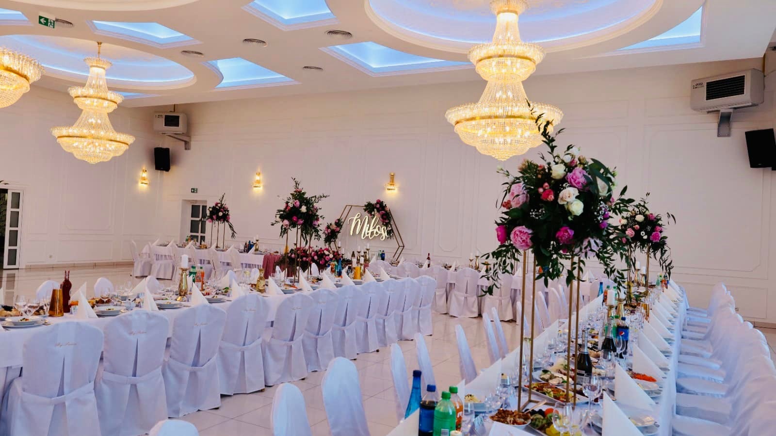 Uroczysta sala bankietowa z długimi stołami nakrytymi białymi obrusami i krzesłami w pokrowcach, dekoracje kwiatowe, kryształowe żyrandole, podświetlany napis 'Miłosz' w geometrycznej ramie.