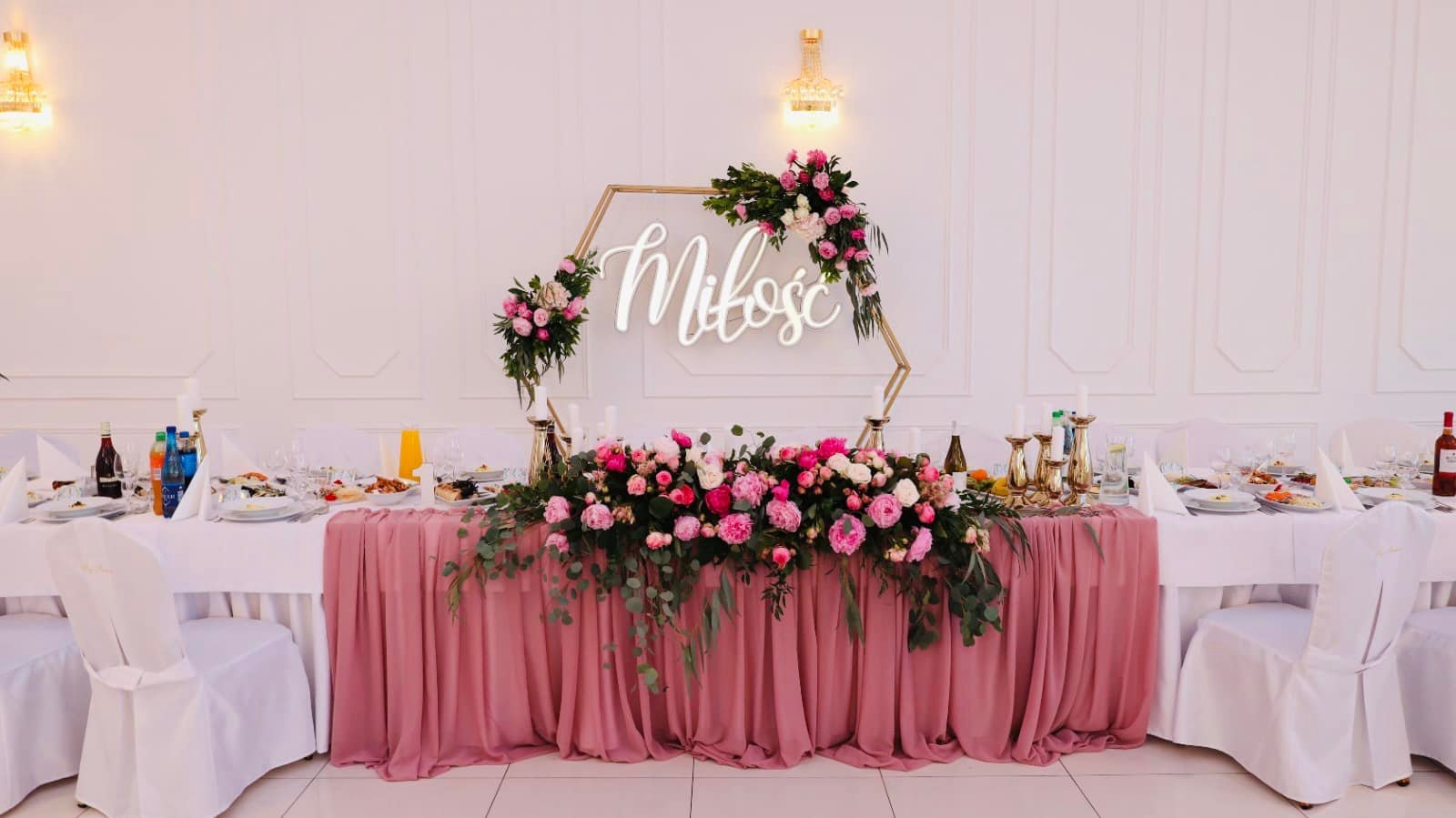 Stół weselny z dekoracją kwiatową w odcieniach różu i bieli, podświetlonym napisem 'Miłość' w geometrycznej ramie, świeczniki i zastawa stołowa na białym obrusie, eleganckie wnętrze sali.