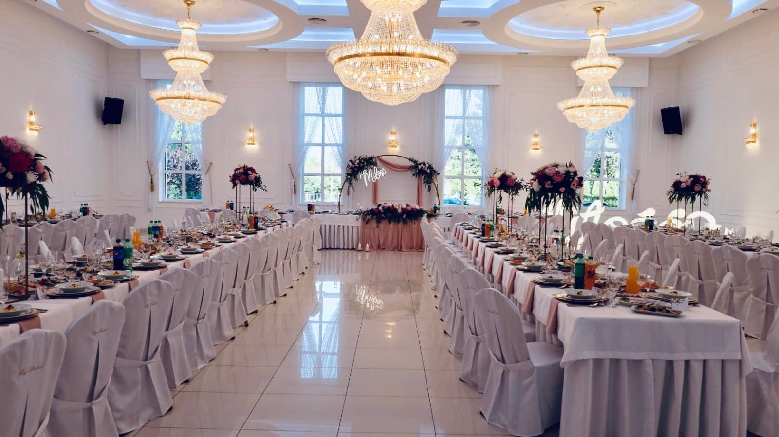 Uroczysta sala bankietowa z długimi stołami nakrytymi białymi obrusami, białymi krzesłami z delikatnymi różowymi akcentami, kwiatowymi dekoracjami i kryształowymi żyrandolami, przygotowana...