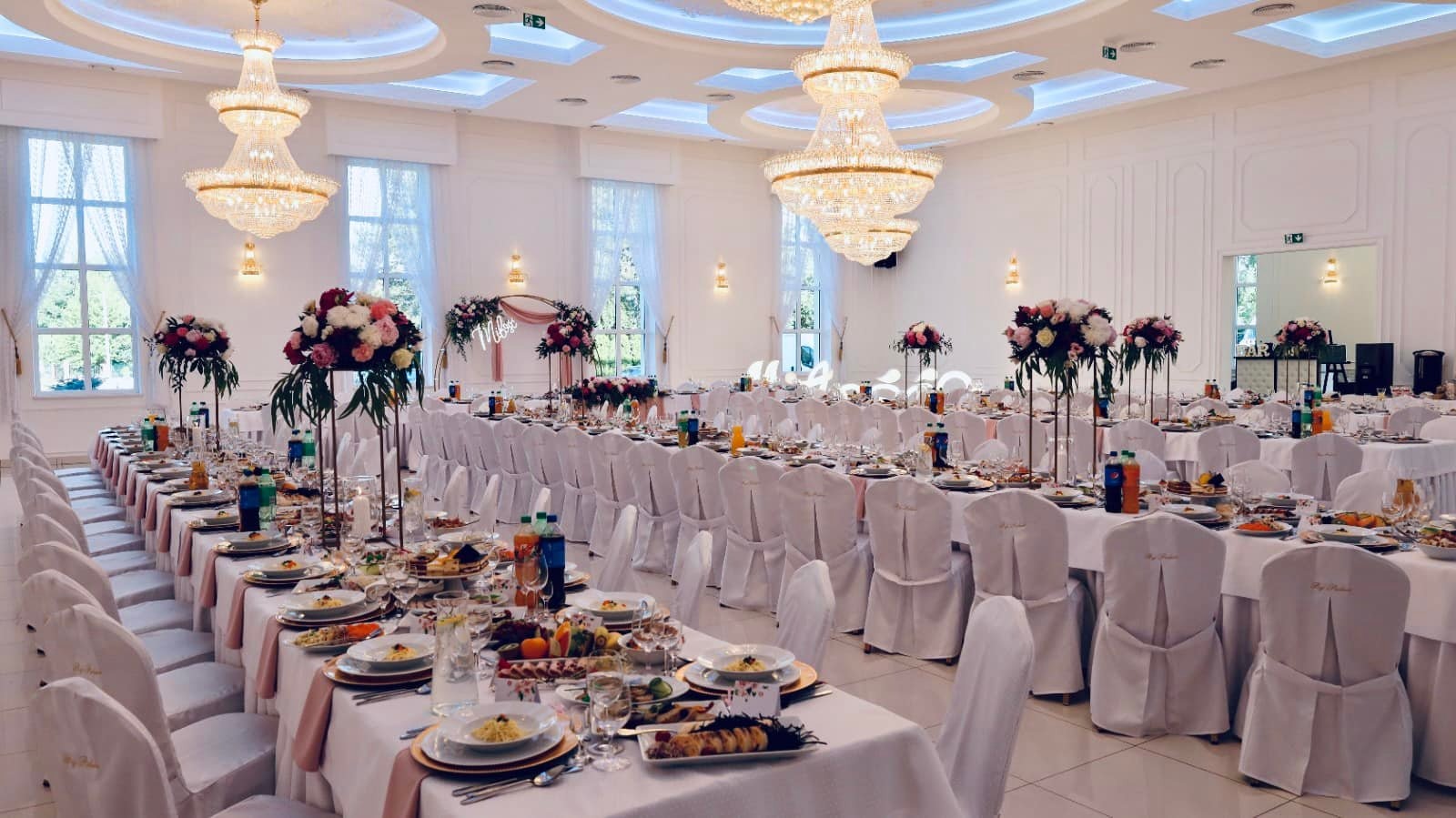 Uroczysta sala bankietowa z długimi stołami nakrytymi białymi obrusami i różowymi bieżnikami, krzesła w białych pokrowcach, wysokie kompozycje kwiatowe, kryształowe żyrandole i dekoracyjny napis...