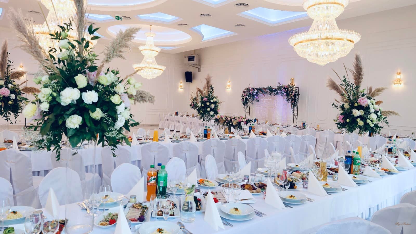 Uroczysta sala bankietowa z długimi stołami nakrytymi białymi obrusami i krzesłami w pokrowcach, bogate kompozycje kwiatowe i wysokie świeczniki, kryształowe żyrandole.