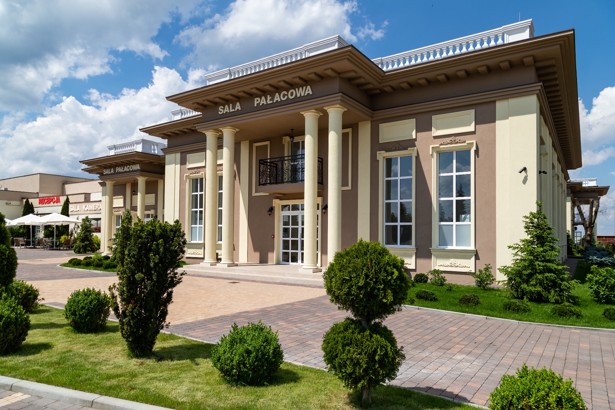 Elewacja dwupiętrowego budynku Sala Pałacowa z kolumnami, balkonem i zadbanym ogrodem z krzewami.