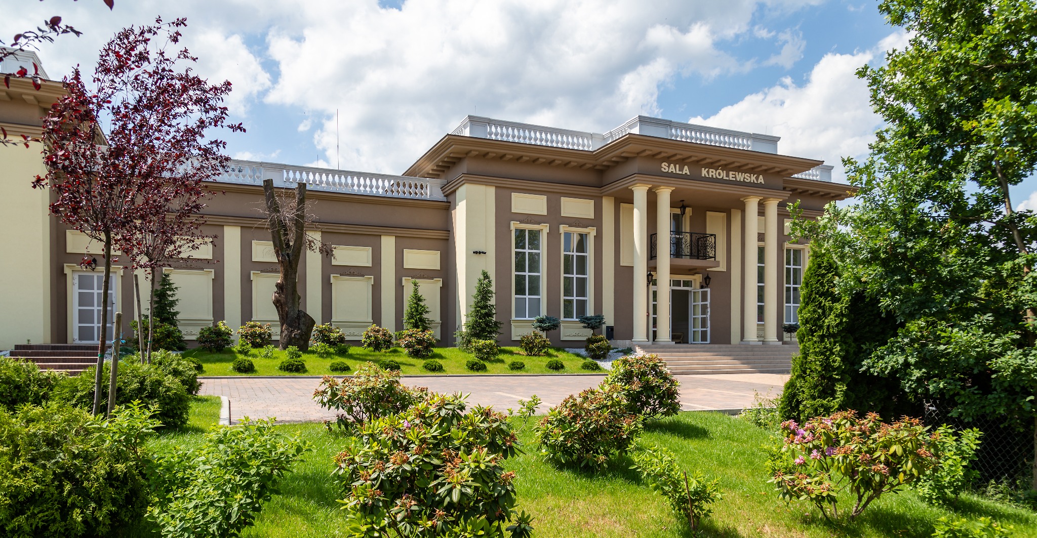 Elewacja budynku Sala Królewska z kolumnami, balkonem i zielonym ogrodem z krzewami i drzewami, widok z zewnątrz w słoneczny dzień.