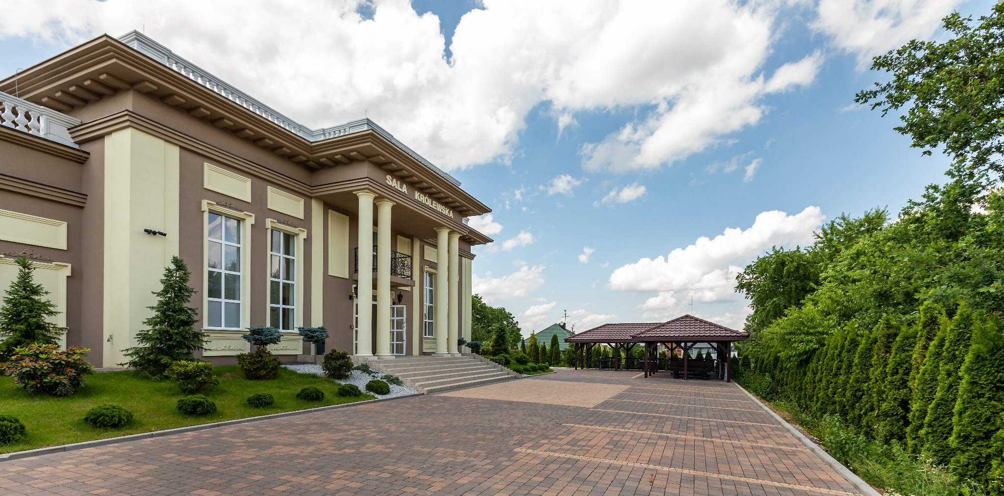 Budynek sali bankietowej Sala Królewska z kolumnami i schodami wejściowymi, widok z zewnątrz na tle błękitnego nieba z chmurami oraz altany ogrodowej.