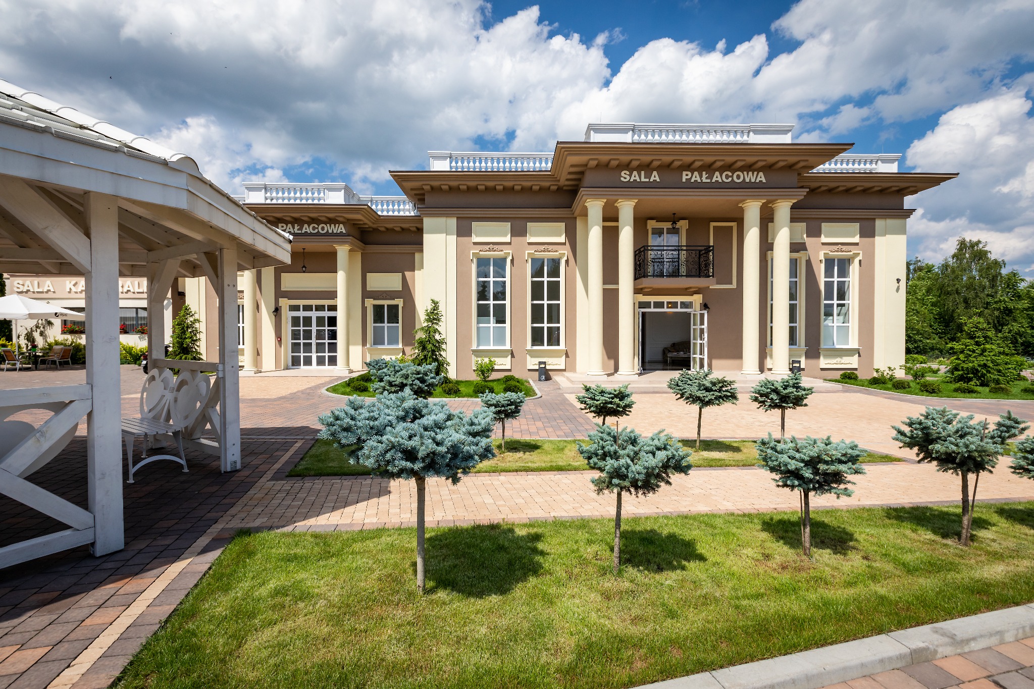Elegancki budynek 'Sala Pałacowa' z kolumnami i balkonem, otoczony zielenią i ozdobnymi drzewkami, widok z zewnątrz w słoneczny dzień.