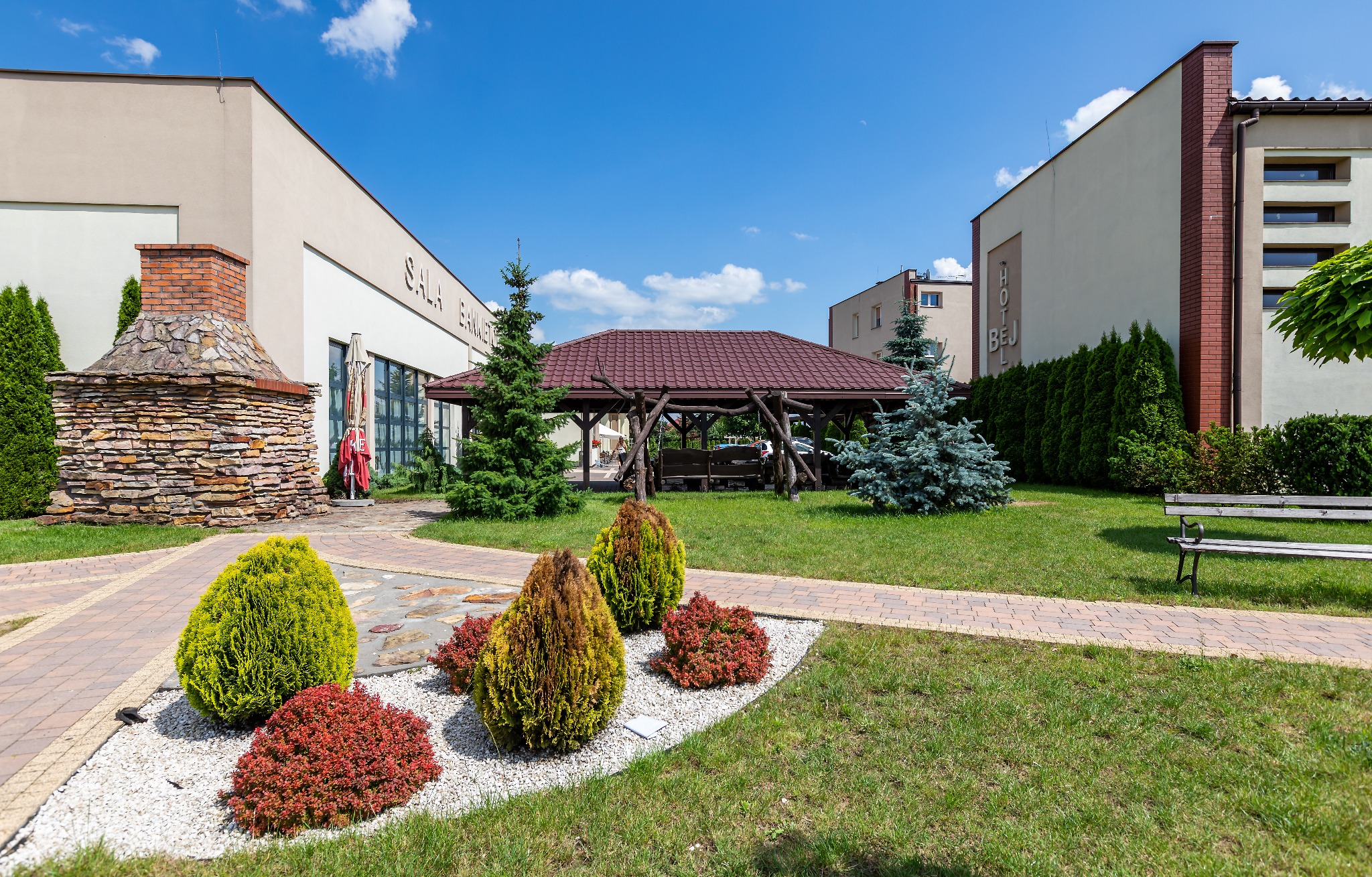 Zewnętrzny widok na salę bankietową z kamiennym kominkiem, altaną z drewnianych bali i zadbanym ogrodem z krzewami i ścieżkami z kostki brukowej.