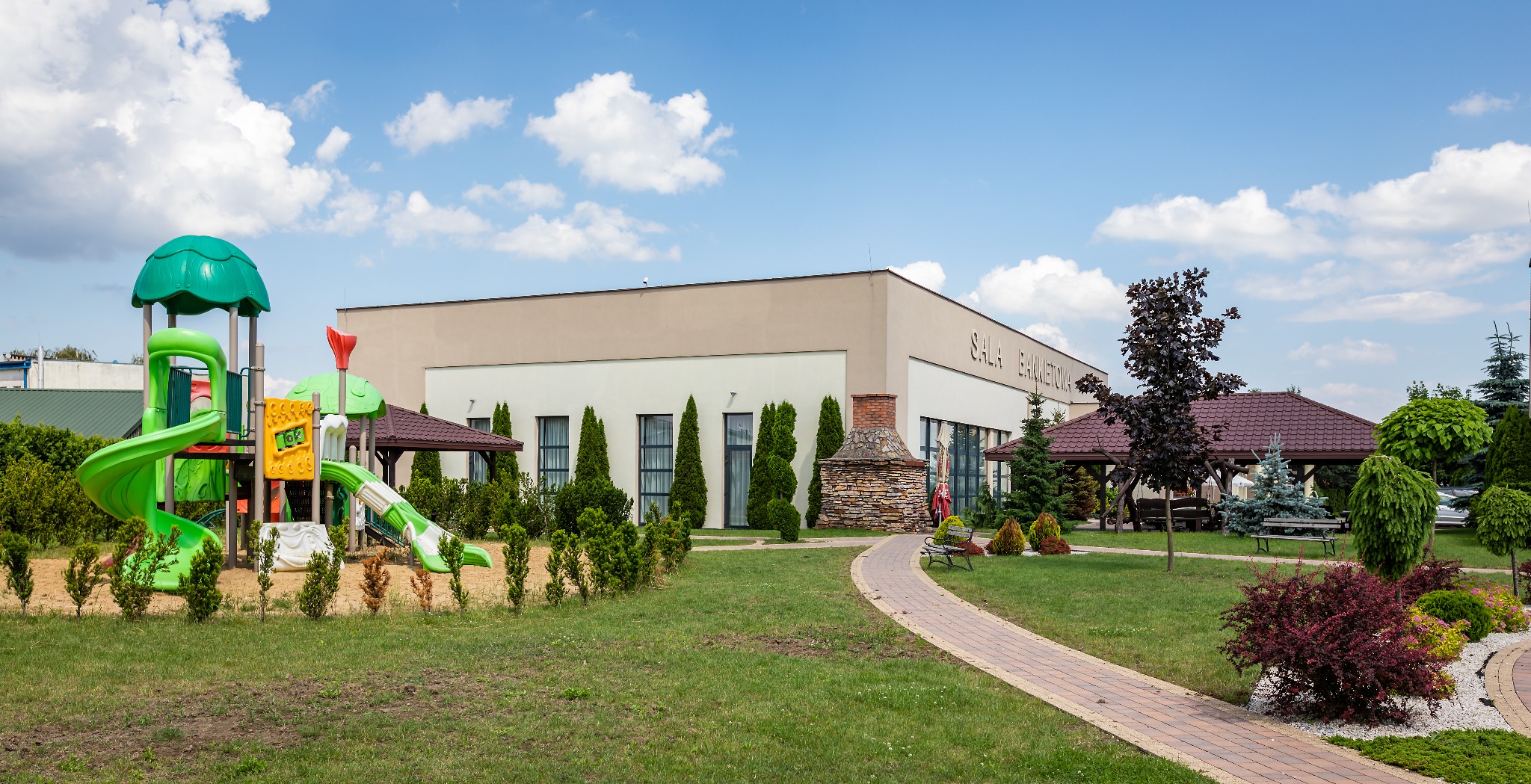 Zewnętrzny widok sali bankietowej z placem zabaw, altanami i zadbanym ogrodem w słoneczny dzień.