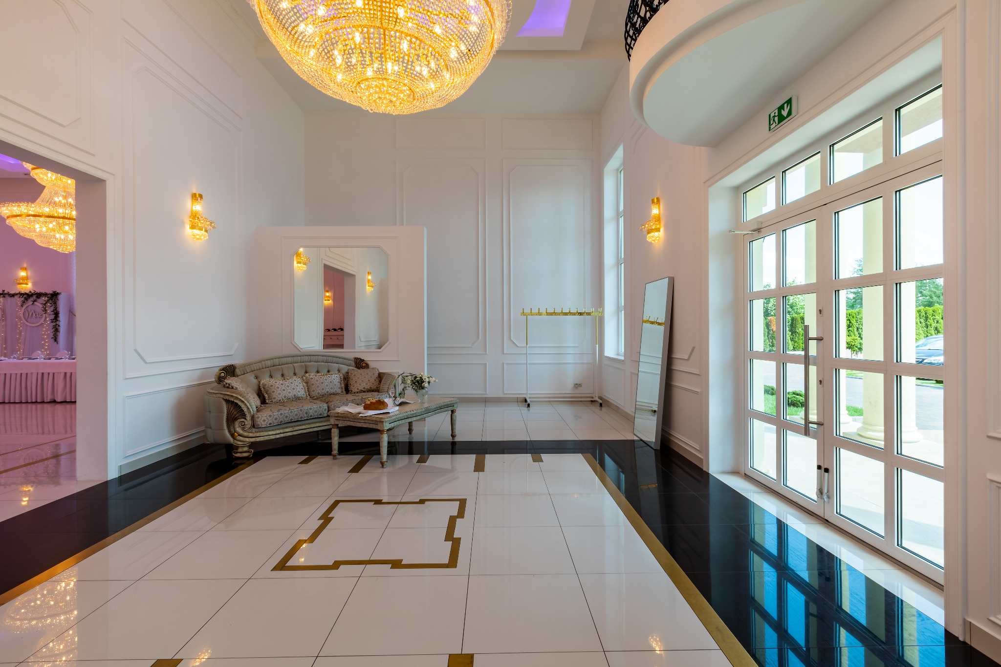 Eleganckie wnętrze sali bankietowej z kryształowym żyrandolem, sofą w stylu vintage i marmurową podłogą w kolorze białym i czarnym ze złotymi akcentami, widok na salę jadalną przez otwarte drzwi.