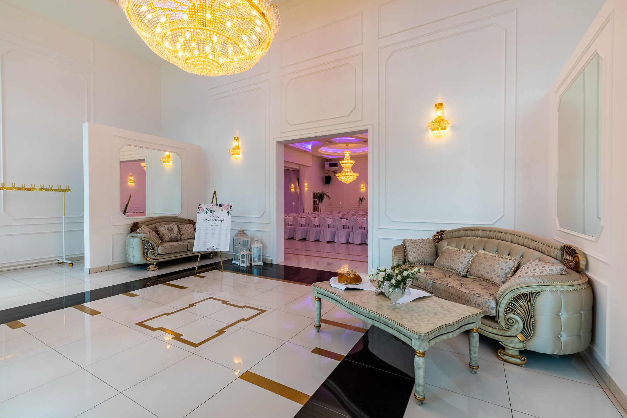 Eleganckie wnętrze sali bankietowej z kryształowym żyrandolem, stolikiem z kwiatami i pieczywem, sofami w stylu vintage oraz widokiem na salę z nakrytymi stołami.