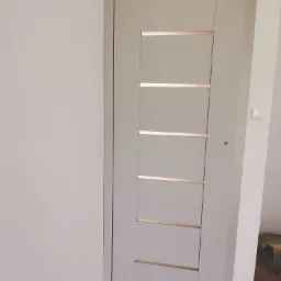 Szare skrzydło drzwiowe z poziomymi, metalowymi wstawkami, zamontowane w jasnym wnętrzu z drewnianą podłogą.