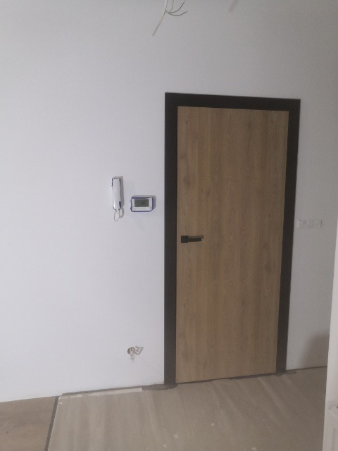 Nowo zamontowane, drewnopodobne skrzydło drzwiowe z ciemną ościeżnicą i czarną klamką, widoczne niedociągnięcia wykończeniowe na ścianie i podłodze.