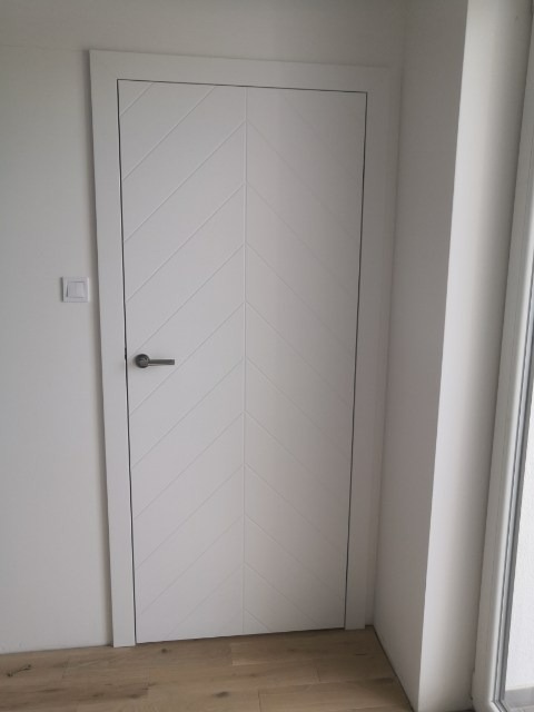 Białe skrzydło z frezowanym wzorem w jodełkę, zamontowane w białej ościeżnicy, widoczne na tle białej ściany i drewnianej podłogi.