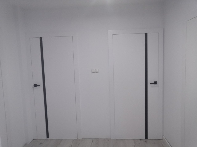 Dwa białe skrzydła z czarnymi, pionowymi intarsjami i czarnymi klamkami na tle białych ścian i jasnej podłogi.