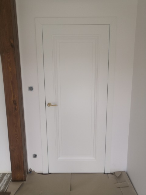 Białe drzwi wewnętrzne z dekoracyjnymi frezowaniami i złotą klamką, zabezpieczone kartonem na podłodze podczas remontu, widoczny fragment drewnianej belki.