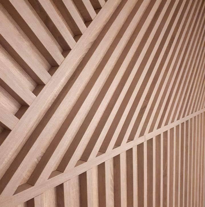 Drewniana ściana z pionowych listew, część z nich ułożona pod kątem tworząc geometryczny wzór, jasne drewno.