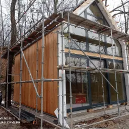 Budowa domu szkieletowego w trakcie wykańczania elewacji drewnianej; widoczne rusztowania, duże okna i las w tle.