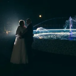 Para młoda w objęciach na tle podświetlanej fontanny nocą, w tle altana i lampy.