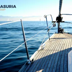 Widok z dziobu jachtu żaglowego na spokojne morze, w tle zarys gór, logo firmy Masuria Group w lewym górnym rogu.