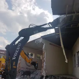 Żółto-czarna koparka Volvo z hydraulicznym młotem pneumatycznym rozbija ścianę budynku z cegieł i pustaków pod błękitnym, zachmurzonym niebem.