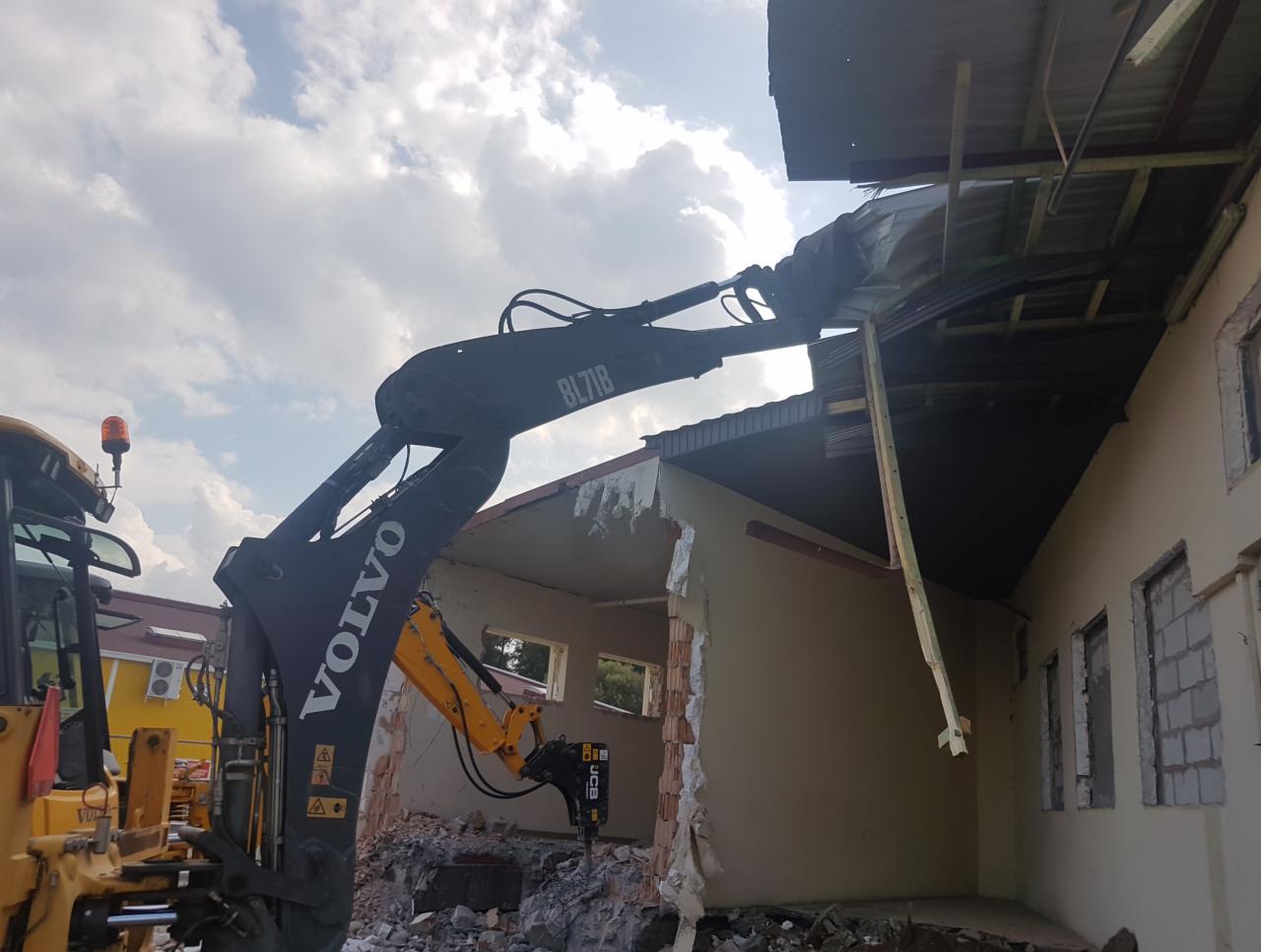 Żółto-czarna koparka Volvo z hydraulicznym młotem pneumatycznym rozbija ścianę budynku z cegieł i pustaków pod błękitnym, zachmurzonym niebem.