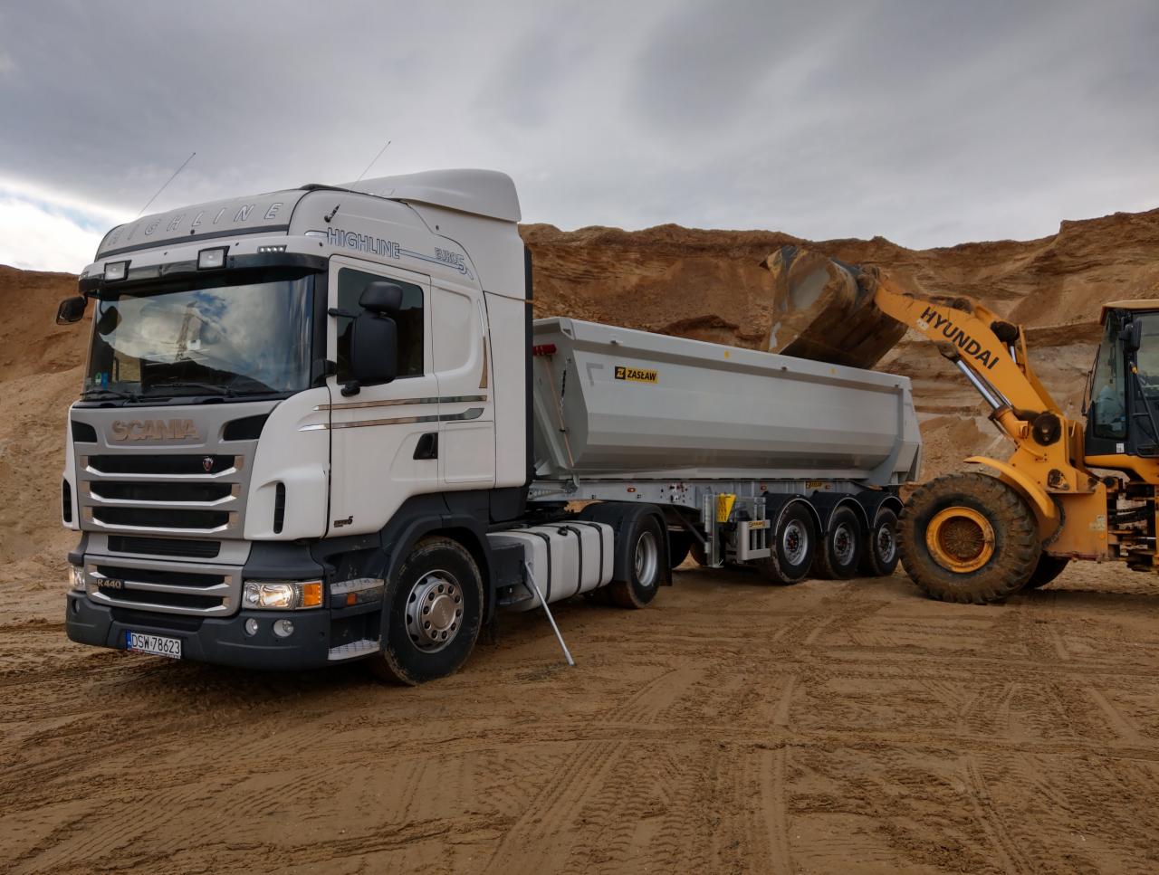 Żółta ładowarka Hyundai na tle piaskowej skarpy, wsypuje urobek do naczepy ciężarówki Scania Highline, stojącej na piaszczystym podłożu pod pochmurnym niebem.