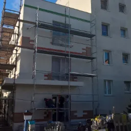 Dwóch pracowników na rusztowaniu przy ocieplanej elewacji bloku mieszkalnego, widoczne fragmenty izolacji, pojemniki z zaprawą i narzędzia na chodniku, błękitne niebo.