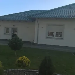 Jasny dom jednorodzinny z ciemną dachówką, widok z ogrodu z ozdobnymi drzewkami iglastymi.