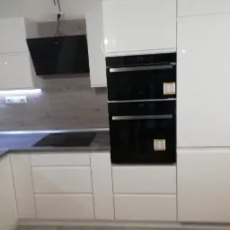 Biała, nowoczesna zabudowa kuchenna z dwoma czarnymi piekarnikami w zabudowie, szarym blatem i drewnopodobną ścianą nad blatem, oświetlona listwą LED pod szafkami.