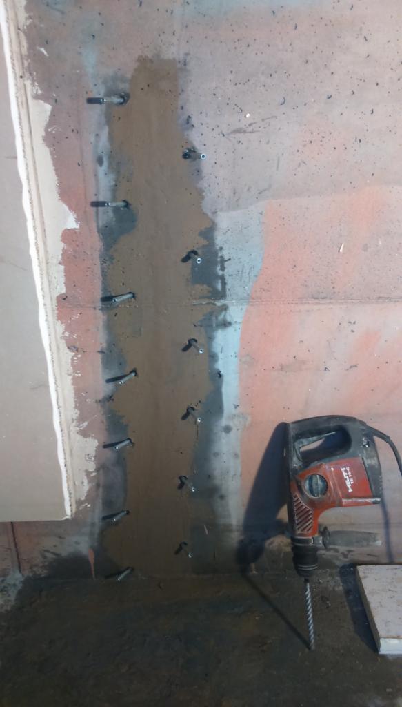 Wzmocnienie strukturalne ściany z widocznymi kotwami i śladami wilgoci, w tle czerwona ściana, obok wiertarka udarowa Hilti TE 16-C.