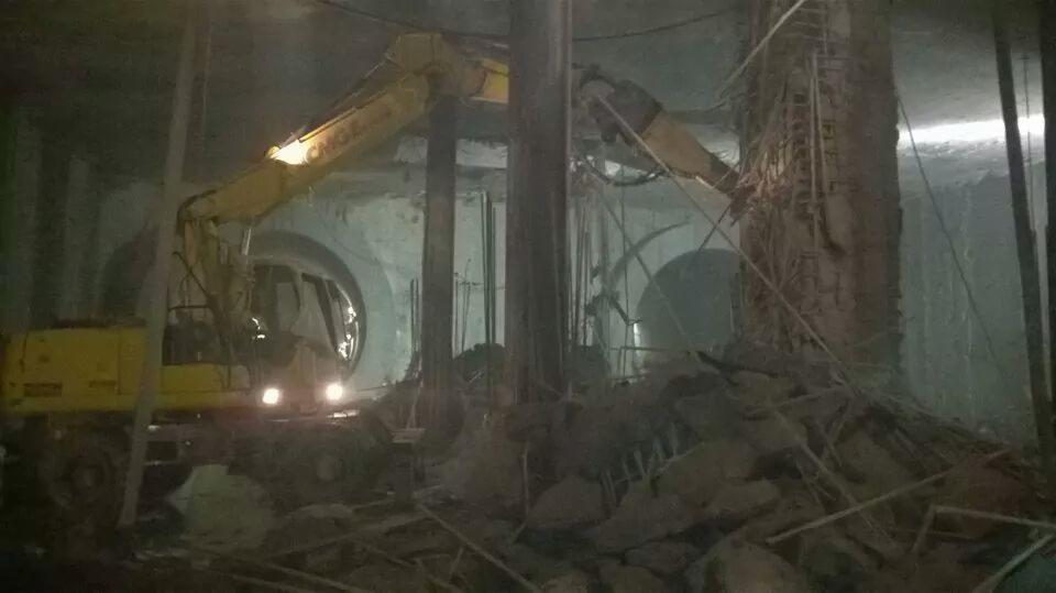 Żółta koparka z pneumatycznym młotem rozbija betonową konstrukcję w tunelu. Wokół gruz i pył.