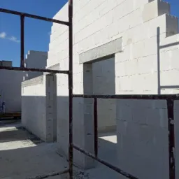 Wznoszenie ścian domu z białych bloczków betonu komórkowego, widoczne otwory okienne i drzwiowe, częściowo zbrojone nadproża, rusztowanie na pierwszym planie.