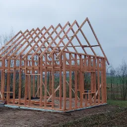 Konstrukcja szkieletowa domu jednorodzinnego w trakcie budowy, widoczne drewniane elementy nośne i podwaliny, przyczepka z piaskiem w tle, pochmurny dzień.