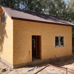 Rubicon montaż - Surowy stan domu szkieletowego z brązowym dachem, widoczne płyty OSB, okno i otwór drzwiowy. Na ziemi narzędzia i elementy konstrukcyjne. Tło: drzewa.