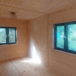 Rubicon montaż - Wnętrze drewnianego domku z dwoma oknami, przez które widać zieleń. Ściany, podłoga i sufit wyłożone jasnym drewnem. Na suficie widoczna lampa z abażurem.