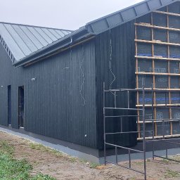 Rubicon montaż - Budowa domu szkieletowego z czarną elewacją i widoczną konstrukcją drewnianą. Na pierwszym planie rusztowanie, w tle dom i przyczepa kempingowa.