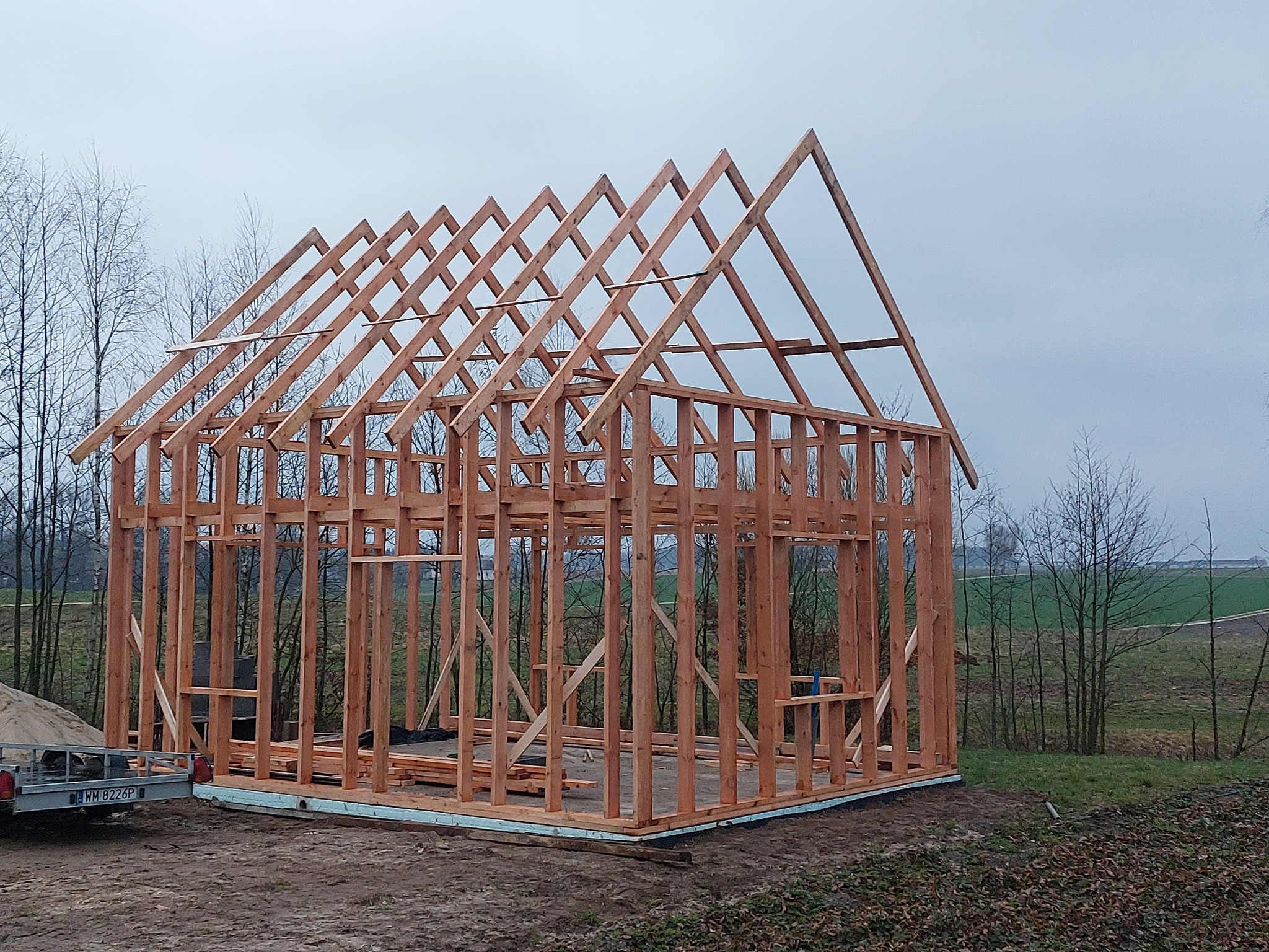 Konstrukcja szkieletowa domu jednorodzinnego w trakcie budowy, widoczne drewniane elementy nośne i podwaliny, przyczepka z piaskiem w tle, pochmurny dzień.