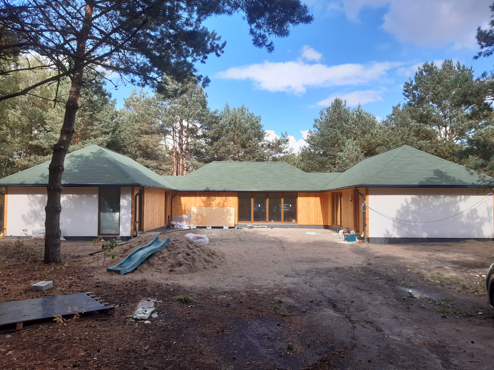 Parterowy dom szkieletowy z zielonym dachem w lesie, częściowo wykończony, z widoczną konstrukcją drewnianą i fragmentami białych ścian. Na podwórku zjeżdżalnia.