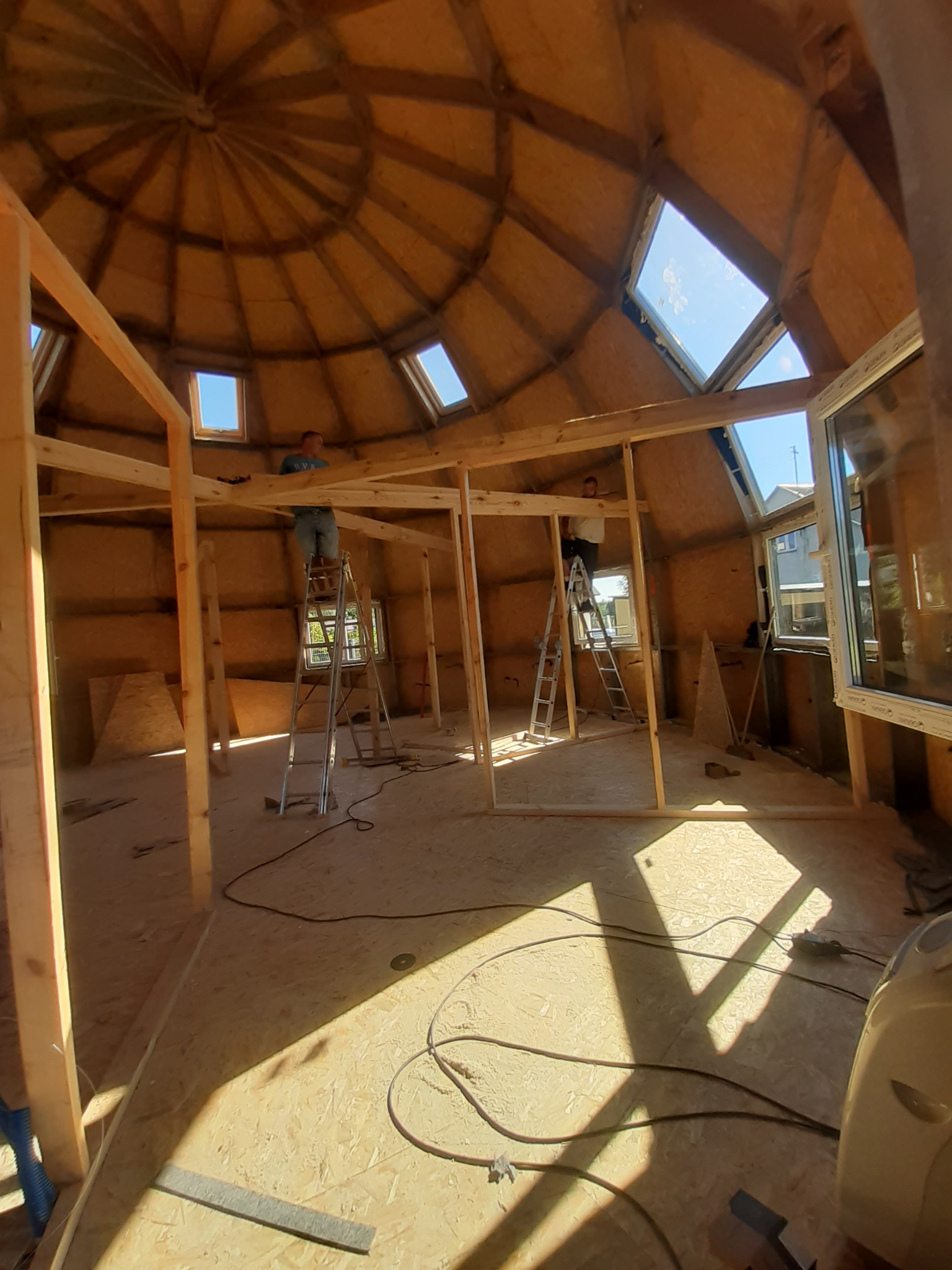 Wnętrze kopuły geodezyjnej w budowie w Mińsku Mazowieckim. Widoczne drewniane ramy, okna i pracownicy na drabinach. Nietypowa konstrukcja dachu.