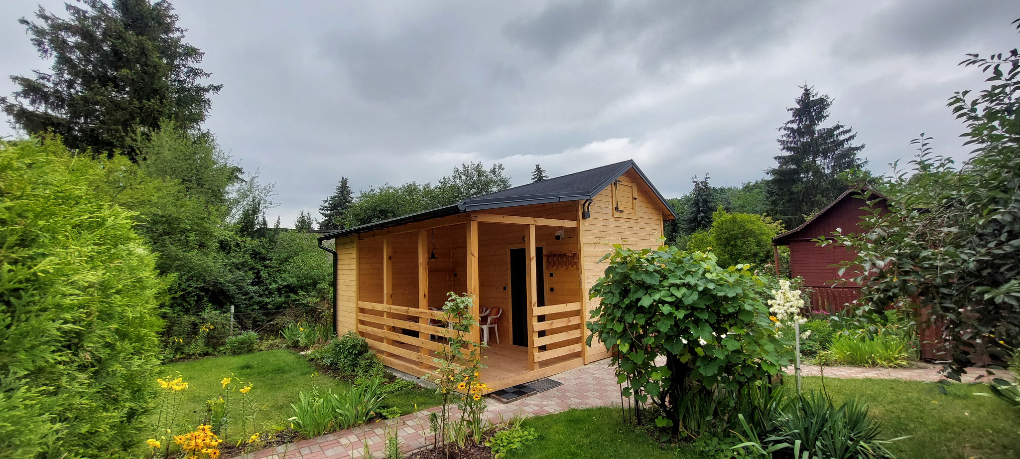 Drewniana altana z zadaszeniem, otoczona zielenią ogrodu, widziana z perspektywy ścieżki. Widoczne kwiaty i krzewy, tworzące naturalne otoczenie.